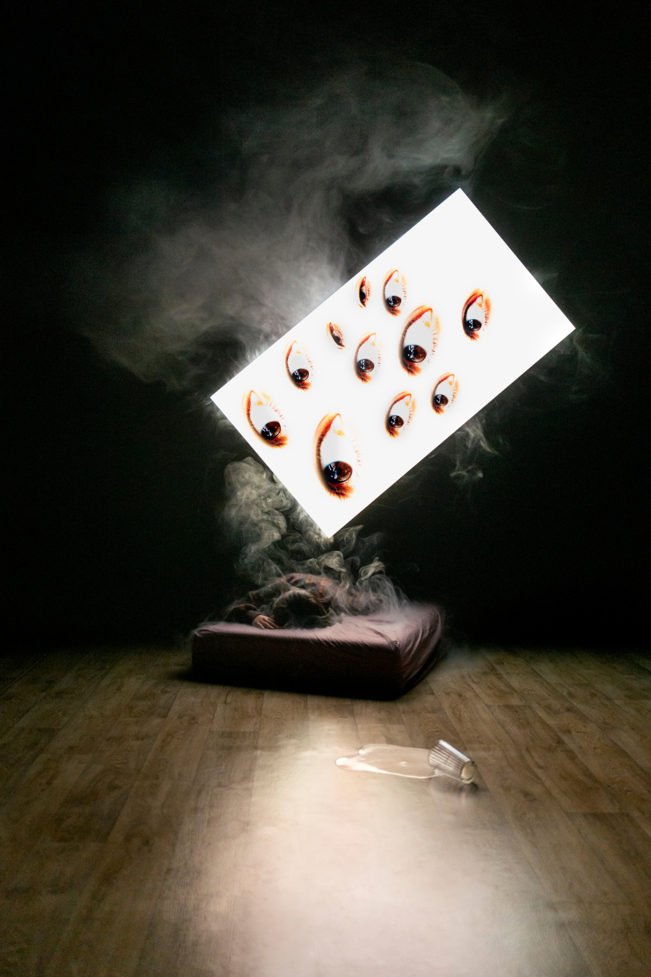 piece of art: a person lying on a mettress surrounded by smoke, a giand card with eyes hovering above